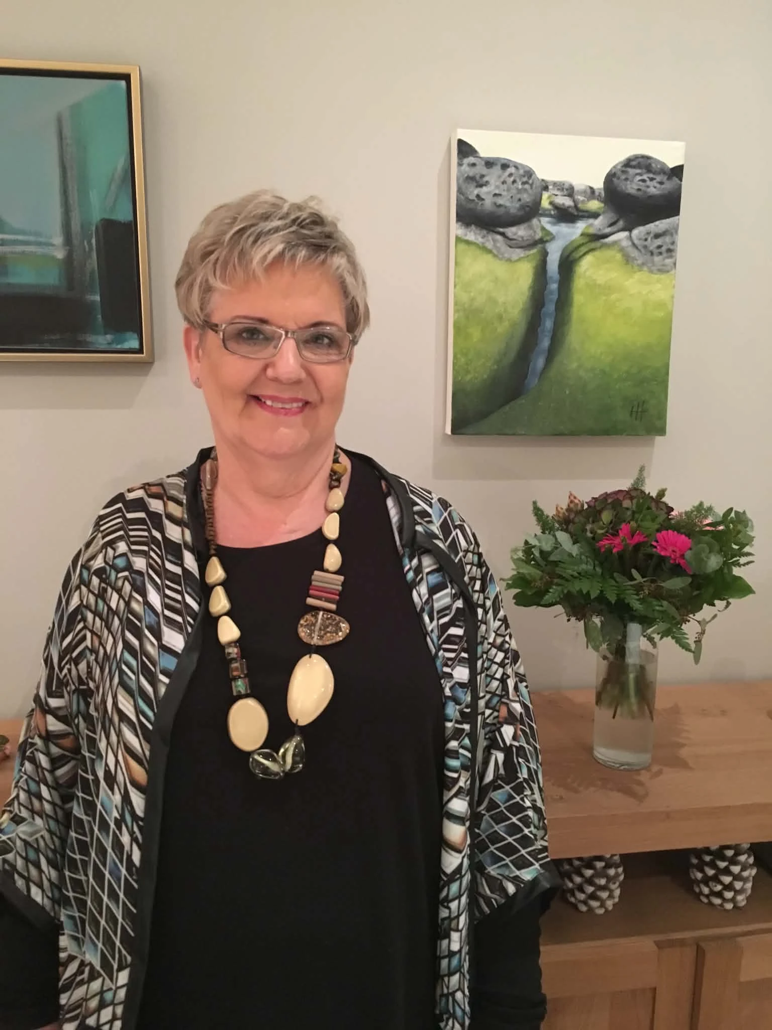 Hrönn Haraldsdóttir stendur við tvö málverk sín, annað af steinum og mosa með læk á milli.